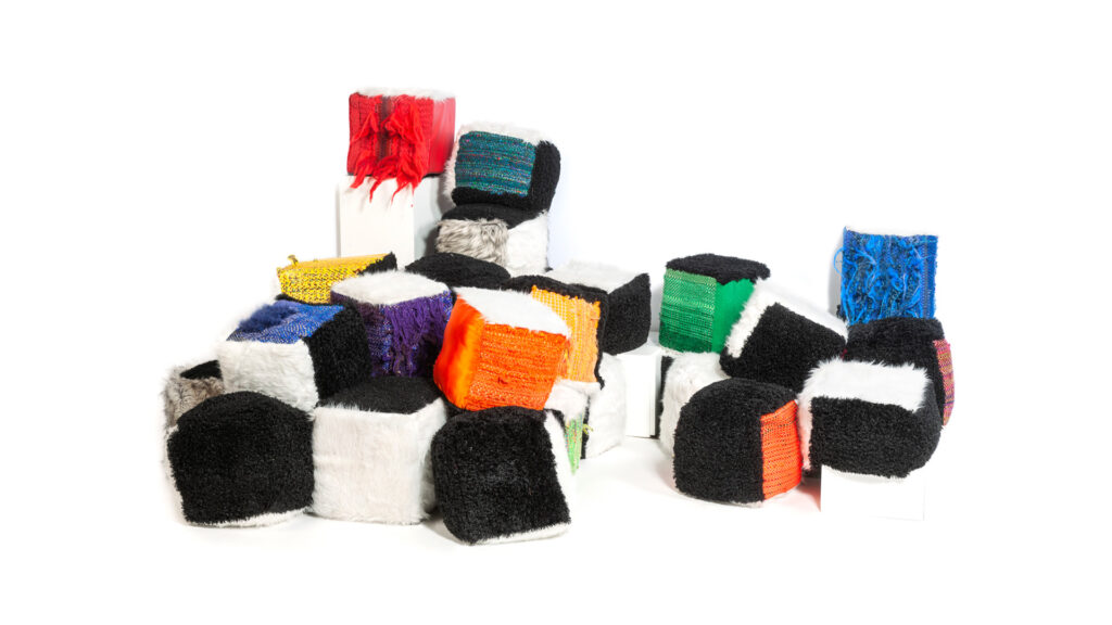 A pile of various textured, cubic soft sculptures on a white background. Some cubes are solid black or white fur, while others are combinations of black, white, orange, red, blue, or green textured fabrics, some with fringe. They are arranged in a loose mound, with a few stacked on white risers.