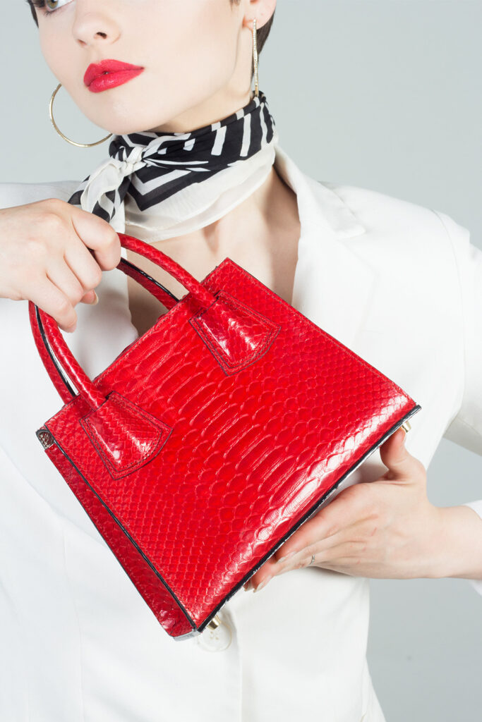 A person from the chest up holds a small, rectangular red purse with a textured, possibly reptilian, surface. They are wearing a white blazer and a black and white patterned scarf. The person has red lipstick and gold hoop earrings.