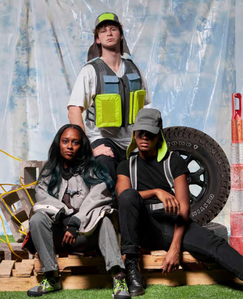Three people are posed in an urban-industrial setting with concrete blocks, pallets, and a large tire. The person in the back wears a white shirt, grey vest with neon yellow pockets, and a matching cap with ear flaps. The person on the left has long, dark hair with green streaks and wears a grey jacket. The person on the right wears a black t-shirt, black pants, and a grey cap with neon yellow accents. All three wear athletic shoes with neon details.