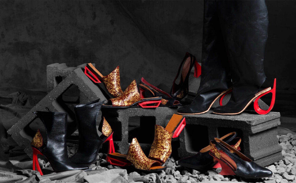 A collection of unique shoes and boots are arranged on and around concrete blocks and gravel against a dark, textured background. Many of the shoes feature black and gold elements with striking red accents, particularly on the heels. One person's legs, clad in black pants and boots, are visible on the right, standing on a concrete block.