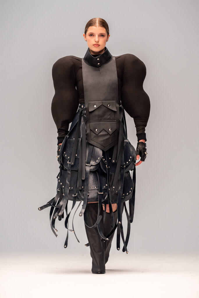 A person walks forward on a light background, wearing a dramatic, all-black ensemble. The outfit features a high-collared top with exaggerated, rounded shoulder pads, a long sleeveless vest adorned with multiple pockets, straps, and buckles, and knee-high boots.