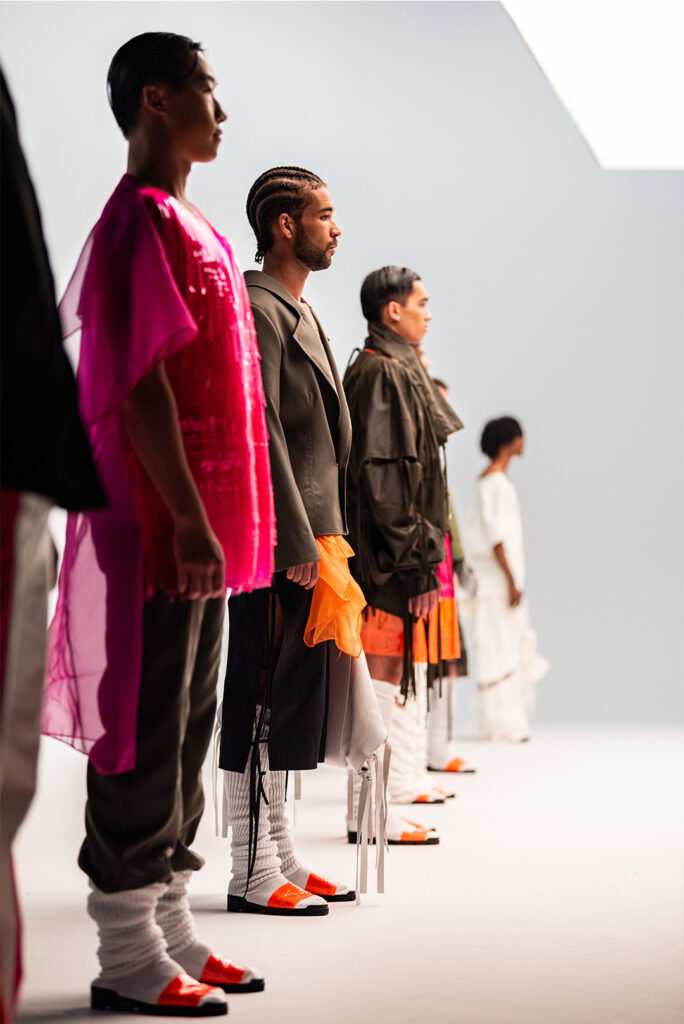 A line of models stands in profile against a light background, showcasing various avant-garde fashion pieces. The closest model wears a sheer, shiny pink top and dark pants with long white leg warmers and orange and black shoes. The other models wear similarly unique and layered outfits with bright accents.