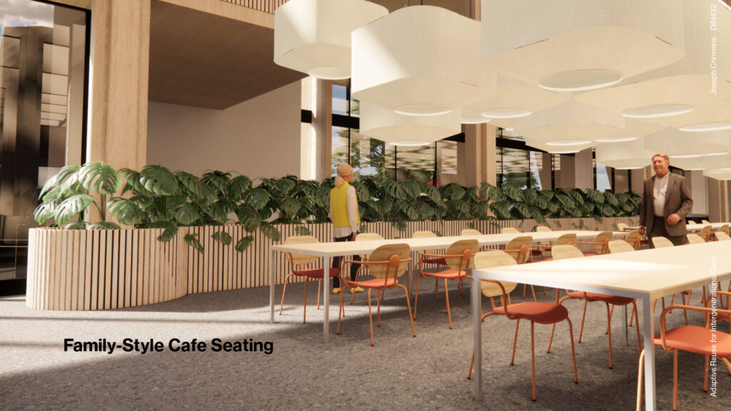 A bright, open family-style cafe seating area with numerous light wood tables and orange chairs. A long planter filled with green plants separates the dining area from a walkway. Large, circular pendant lights hang from the ceiling. People are casually walking or standing in the space.