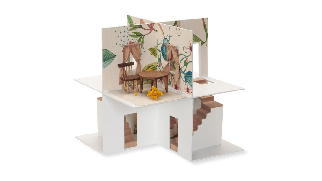 A small, four-sided miniature room model. Two walls are white with doorways, and two walls have a floral wallpaper pattern with windows and draped curtains. A tiny wooden table, two chairs, and a small bunch of yellow flowers are on the floor. A small staircase is built into one side.