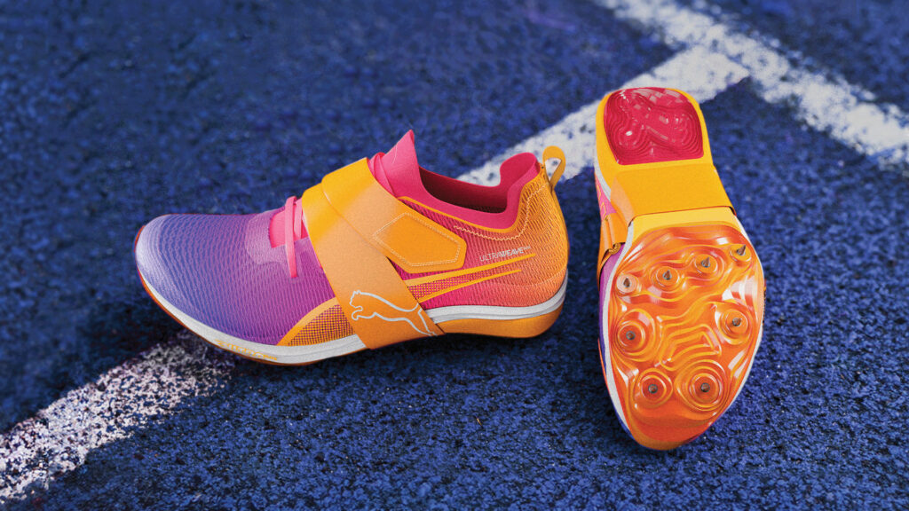 A vibrant, close-up shot of two Puma track and field shoes in shades of purple, pink, and orange, with gold accents. One shoe is seen from the side, and the other from the bottom, showcasing its bright orange spiked sole, both resting on a blue running track with white lines.