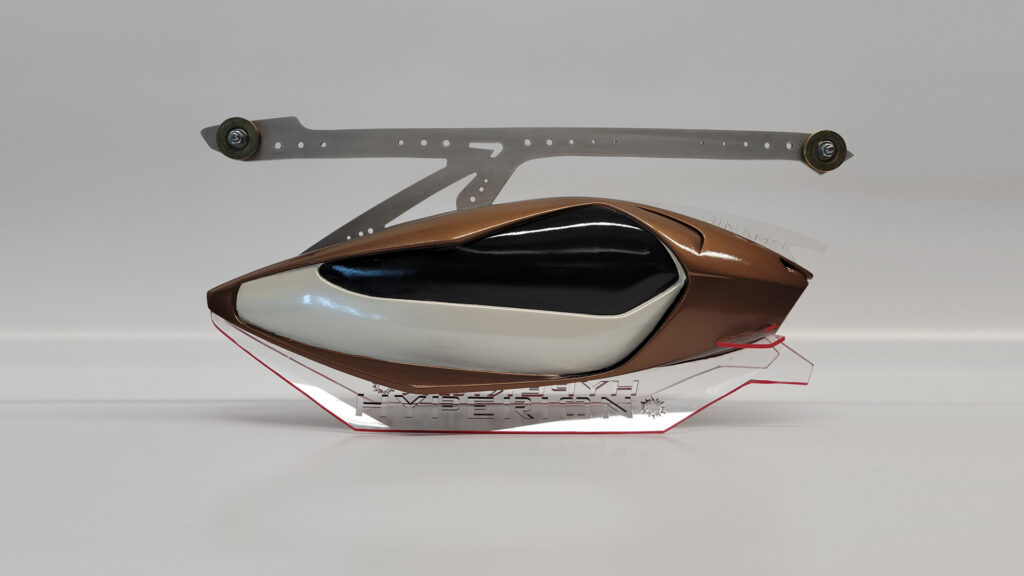 A physical model of a sleek, futuristic vehicle or drone. It is bronze and white with a dark, reflective cockpit area. The model is displayed on a clear, angular base that has text and gear-like graphics, and is supported by a metallic bracket above. The background is plain white.