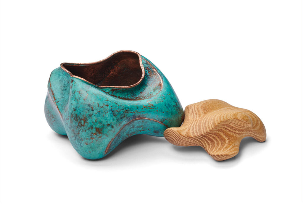 A sculptural piece with two parts on a white background. The main part is an organic, rounded form in a mottled blue-green patina, revealing a coppery interior. Beside it, a separate, intricately carved wooden piece with flowing lines, suggesting a lid or companion piece, rests on the surface.
