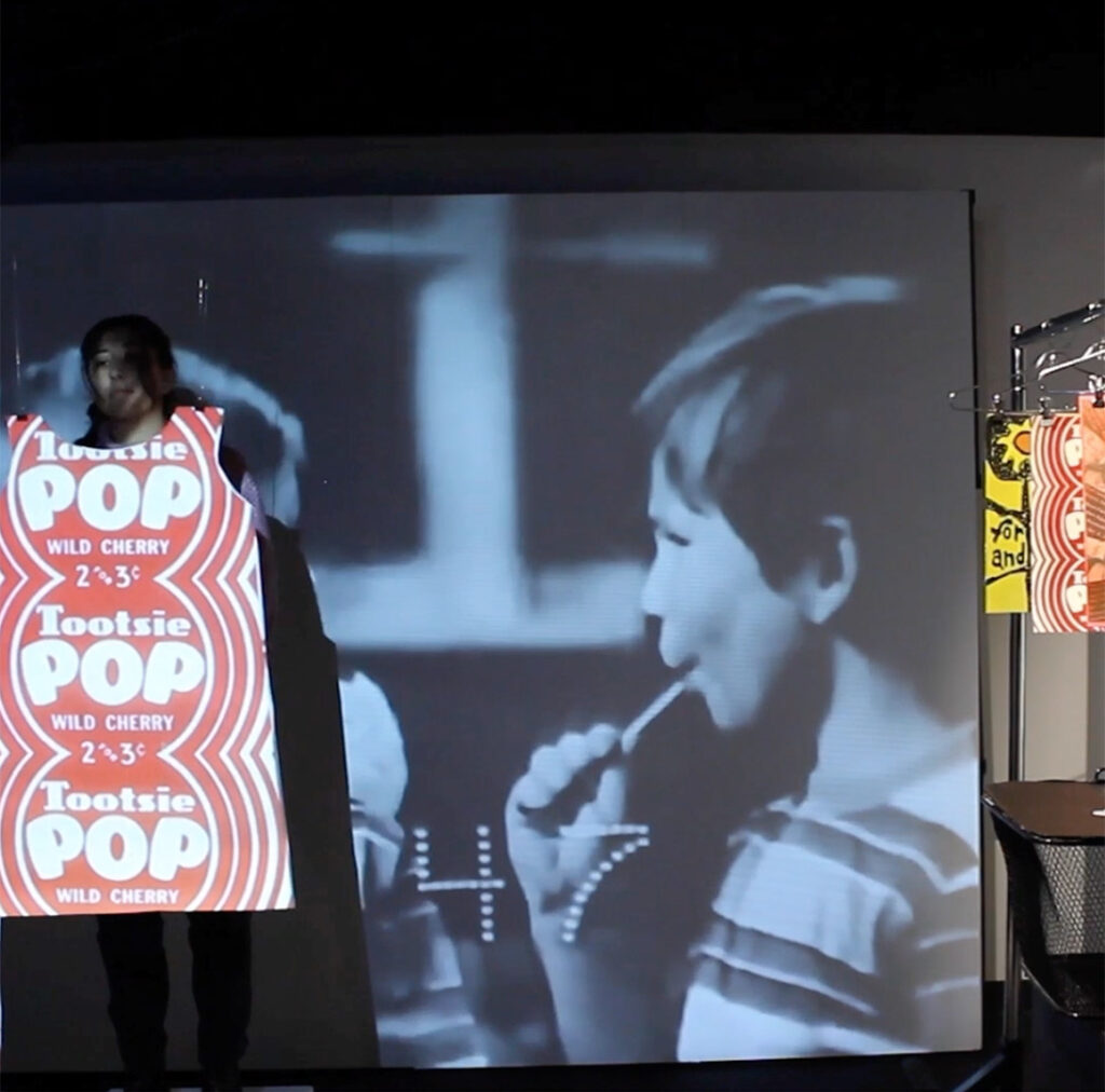 A person stands in front of a large projection screen, wearing a costume designed to look like a giant Tootsie Pop wrapper. The black and white projection behind them shows a young boy sucking on a lollipop. To the right, additional poster-like artworks are visible on a rack.