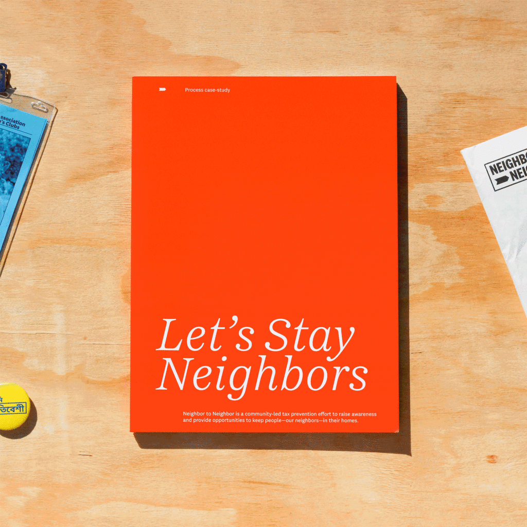 A top-down shot of an orange booklet with the title "Let's Stay Neighbors" in white text, centered on a light wooden surface. Below the title, smaller text describes Neighbor to Neighbor as a community-led tax prevention effort. To the left, a blue ID badge is partially visible, and to the right, a white envelope with the word "NEIGHBO" is seen. The gif shows the booklet slightly opening to reveal blank pages.