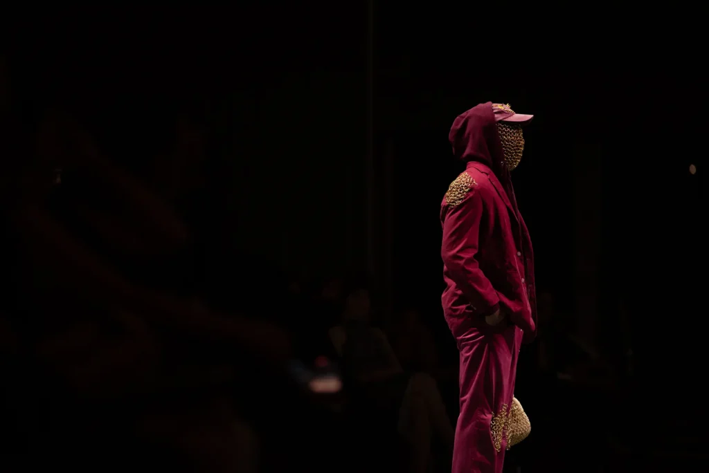A profile view of a model on a runway wearing a deep burgundy-magenta suit, a matching hoodie, and a gold chainmail face mask and shoulder detailing against a dark background.