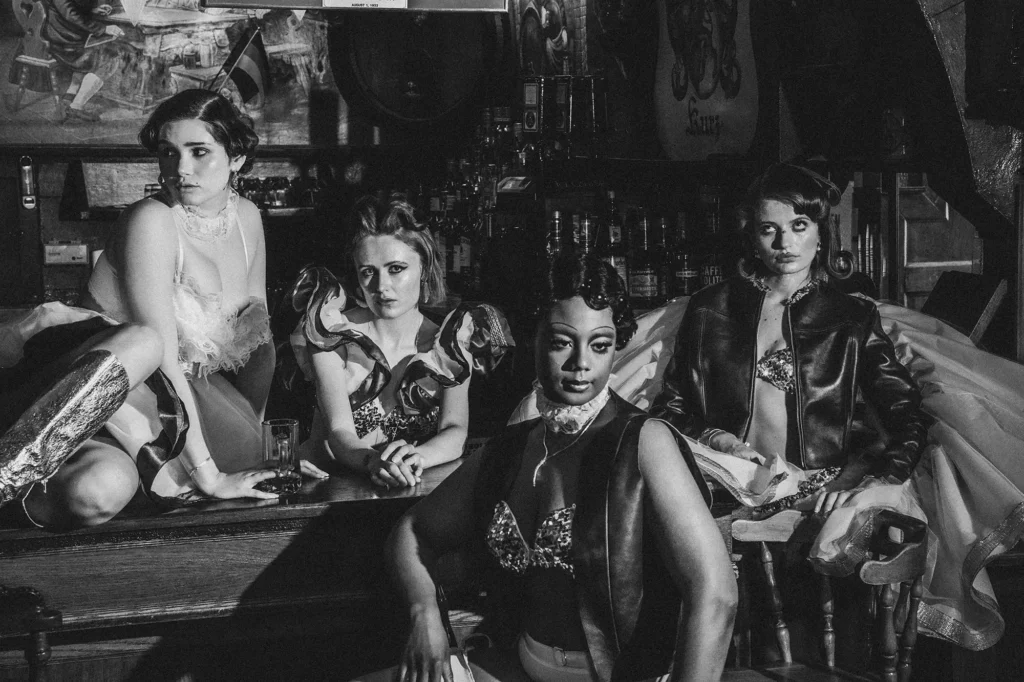 A black-and-white group portrait of four fashion models styled in vintage-inspired garments, posed together in a dimly lit tavern.