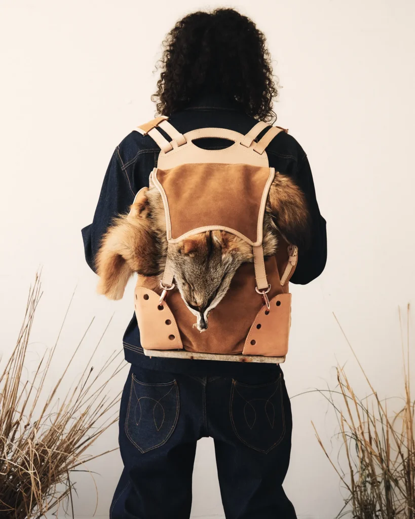 A person from behind wearing dark denim and a tan leather backpack featuring textured animal fur panels and light wood accents.