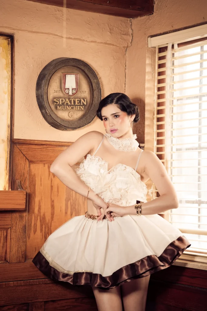 A model posing in a vintage-style tavern wearing a cream-colored lace corset top, a ruffled high collar, and a matching full skirt with a dark brown hem.
